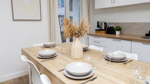 A modern kitchen with white cabinets, a wooden dining table set with plates, glasses, and cutlery, and a decorative vase with pampas grass.