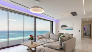 Een moderne woonkamer met een grote schuifglazen deur die leidt naar een balkon met uitzicht op zee. Er staat een beige bank, een houten salontafel en een vaas met bloemen.