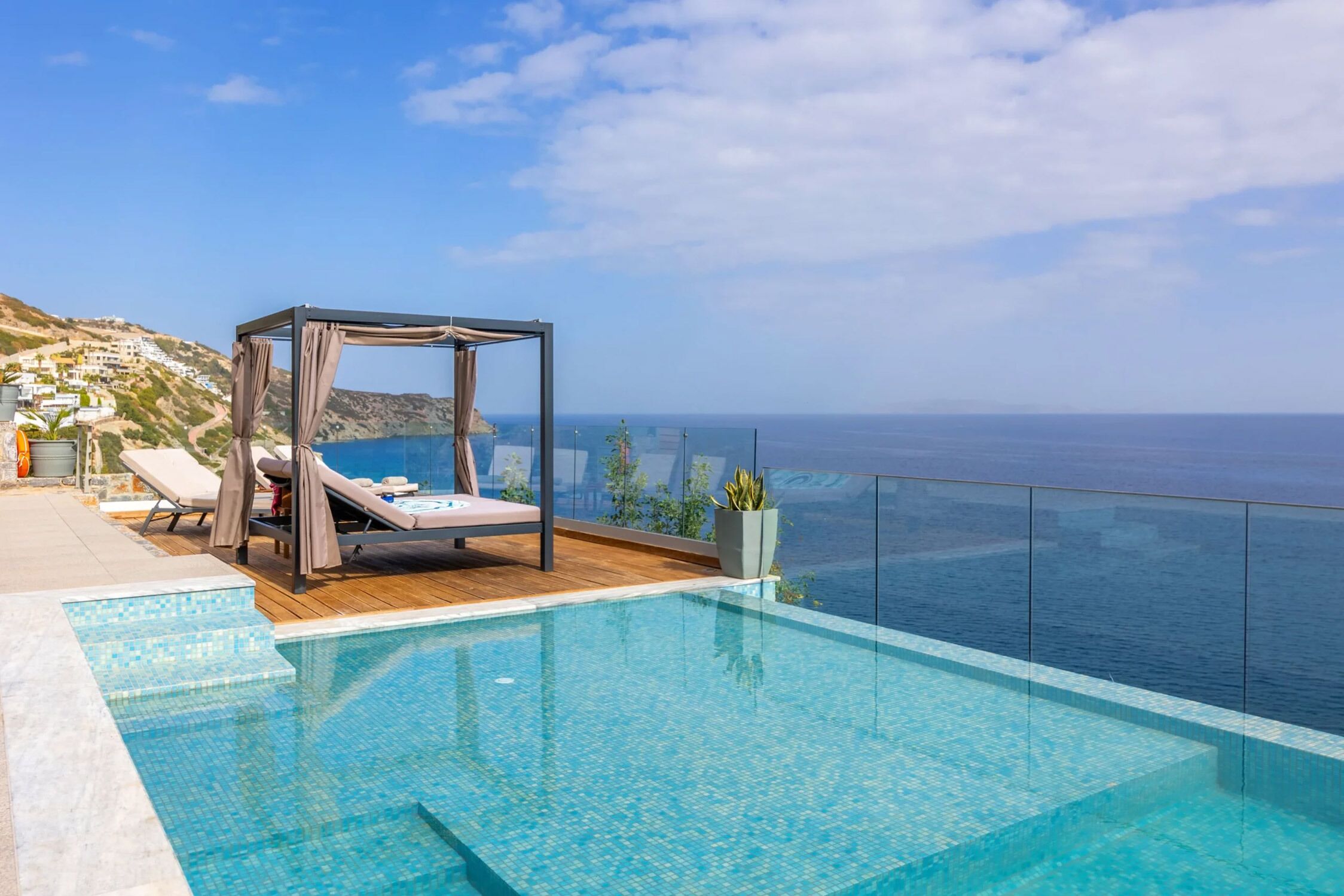 Una piscina infinita moderna con terraza de madera, una cama con dosel y vista despejada al mar.