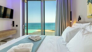 Een hotelkamer met een bed, een aan de muur bevestigde televisie, uitzicht op de oceaan door een schuifdeur en een houten bank.