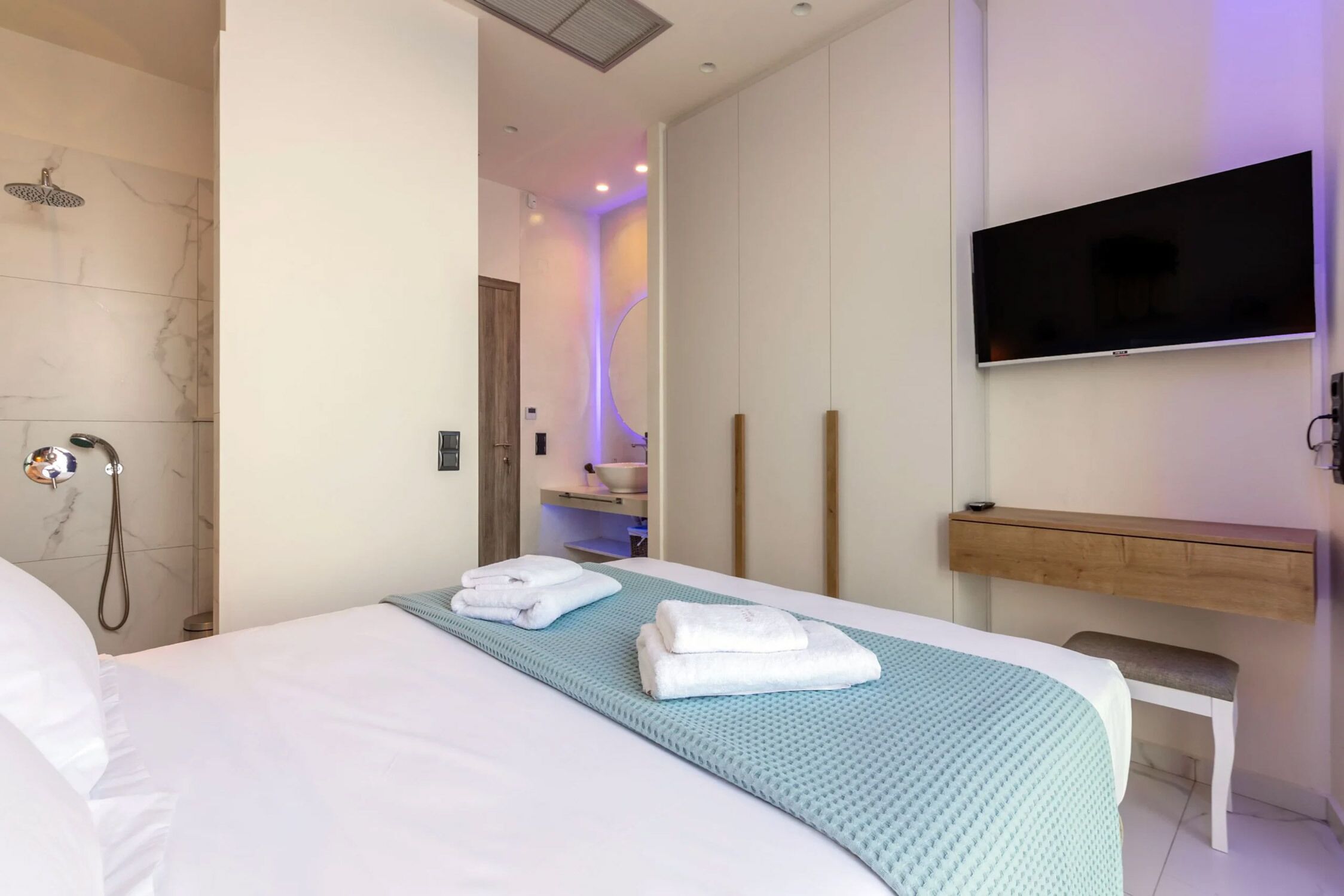 Habitación de hotel moderna con una cama grande, un televisor montado en la pared, un banco de madera y zona de ducha.