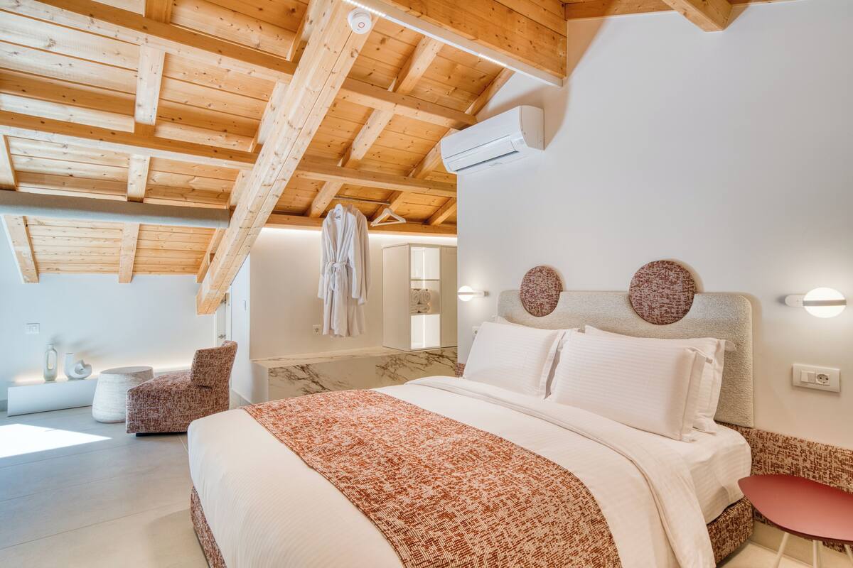 Un dormitorio con techo de madera, una cama con ropa blanca, una silla roja y una mesita.