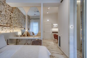A modern hotel room with a marble floor, a large bed, a seating area with a sofa and chairs, a dining area with a table and chairs, and a kitchen visible through a glass door. - Nalia Suites (Nafplio)