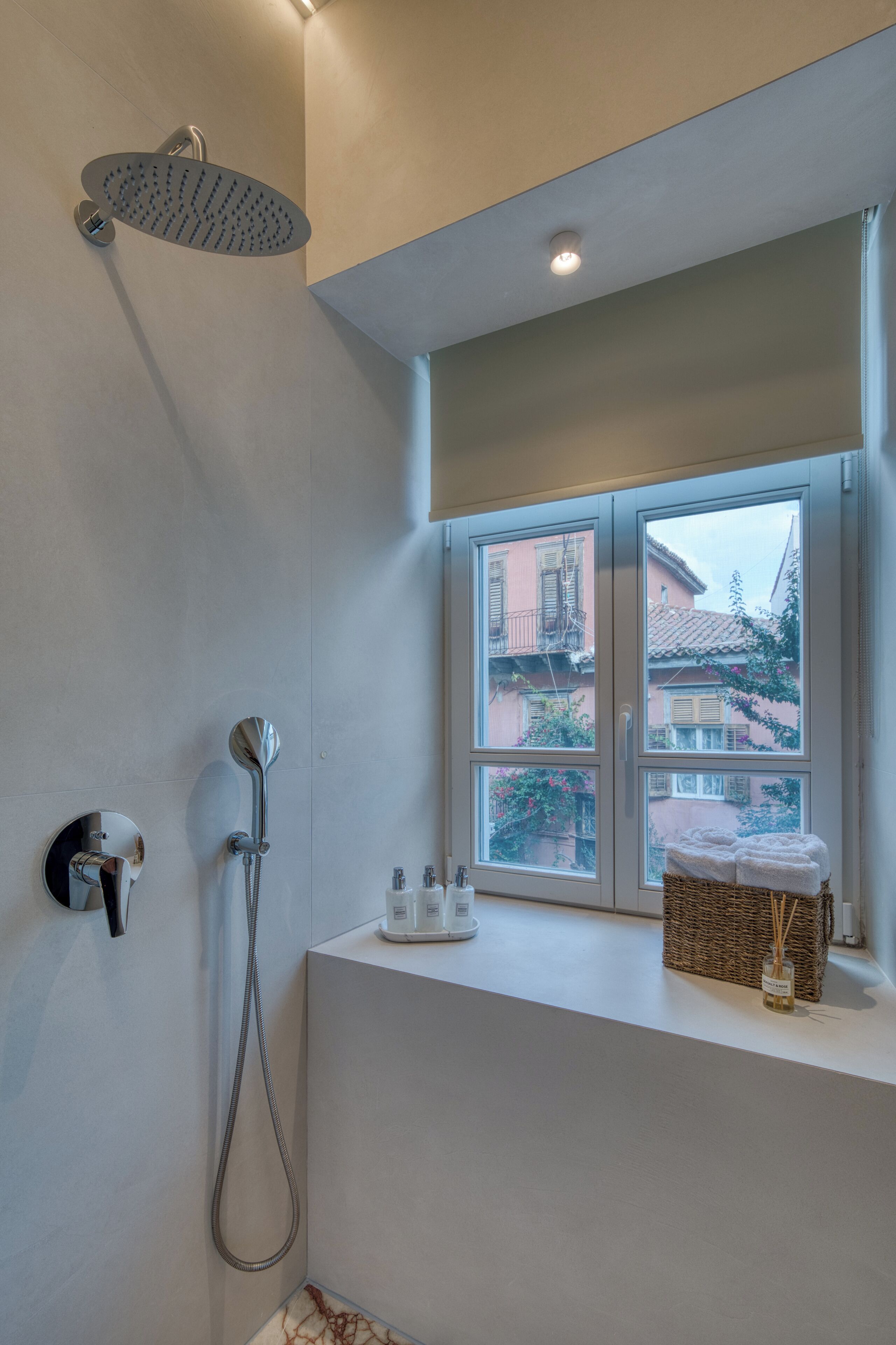 A bathroom with a shower, a window with a view of buildings, and a small shelf with toiletries.