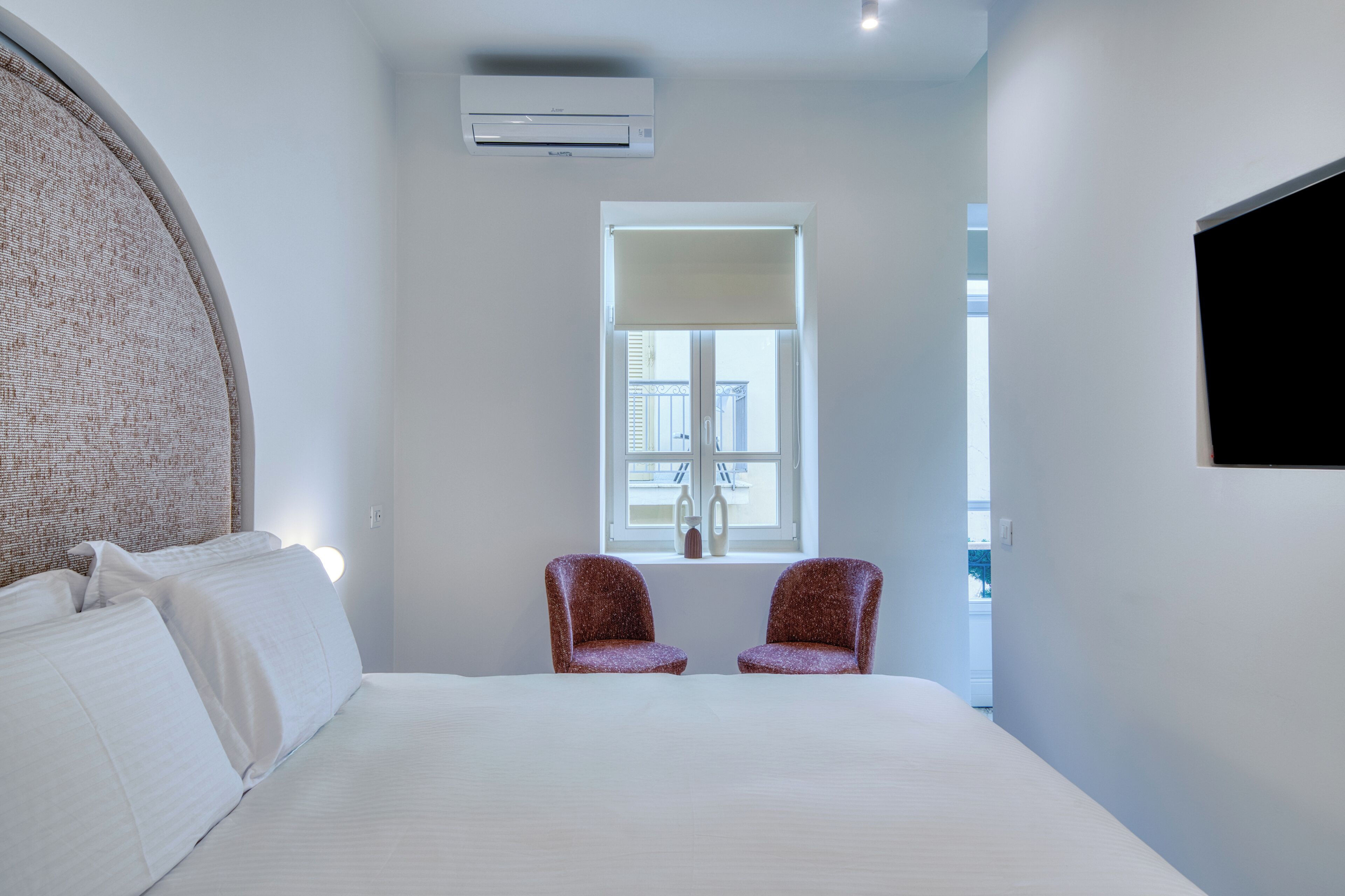 A modern hotel room with a large bed, two armchairs, a window with a view, and an air conditioning unit.