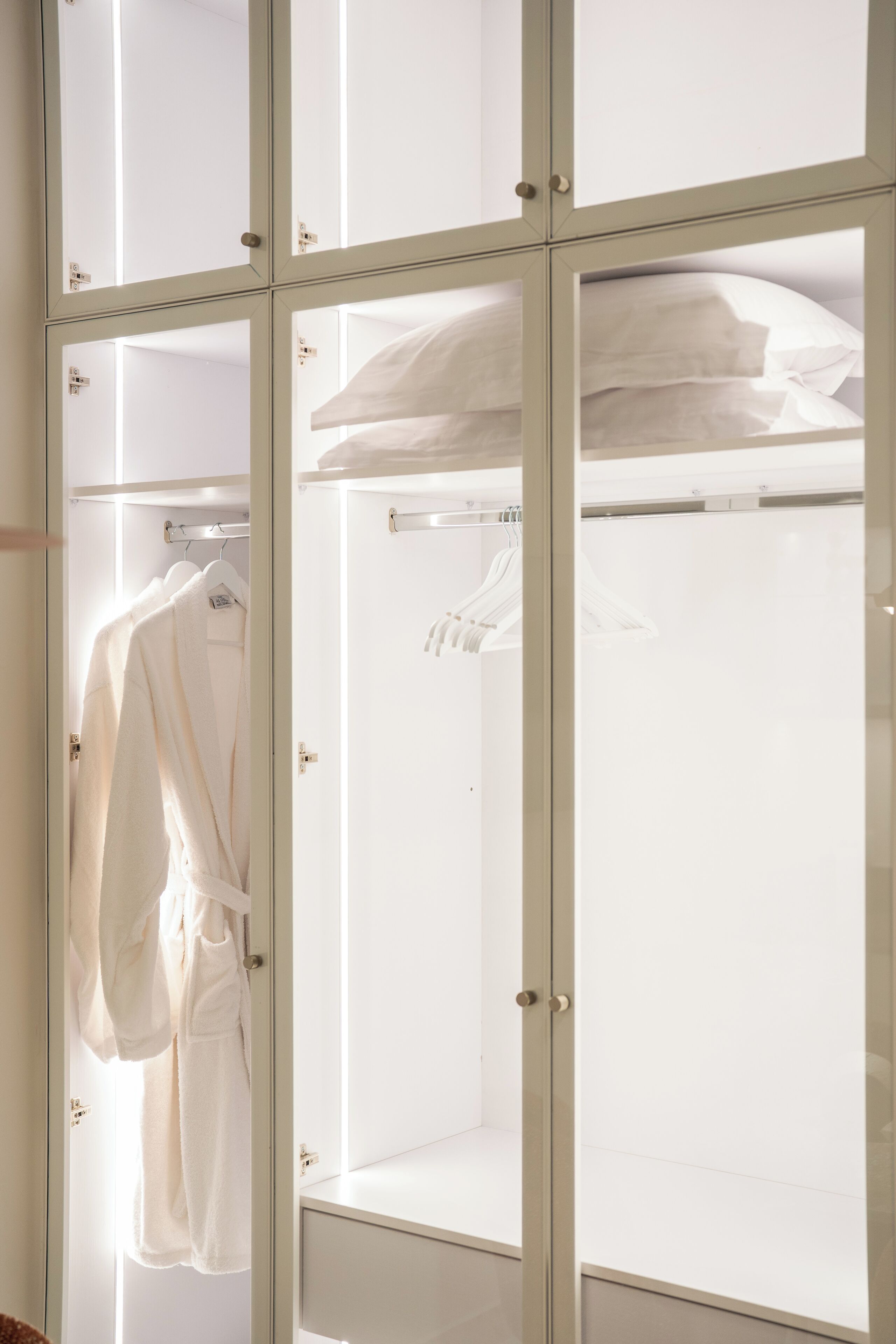 Una habitación con un separador de vidrio, una bata blanca colgada en un perchero y una cama con ropa de cama blanca.