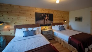 A cozy room with two beds, wooden walls, a mounted painting, and a nightstand with a lamp.