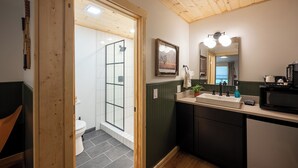 Une salle de bain avec un plafond en bois, des toilettes blanches, une douche avec une porte en verre, un lavabo avec un miroir et une petite plante posée sur le comptoir.