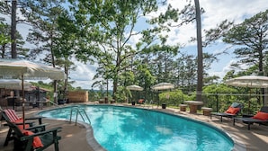 Une piscine entourée d’arbres et d’une clôture, avec des chaises longues et des parasols.