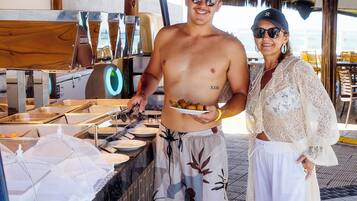 Un hombre y una mujer están de pie cerca de una mesa de buffet con suelo de baldosas y una estructura de techo de paja.