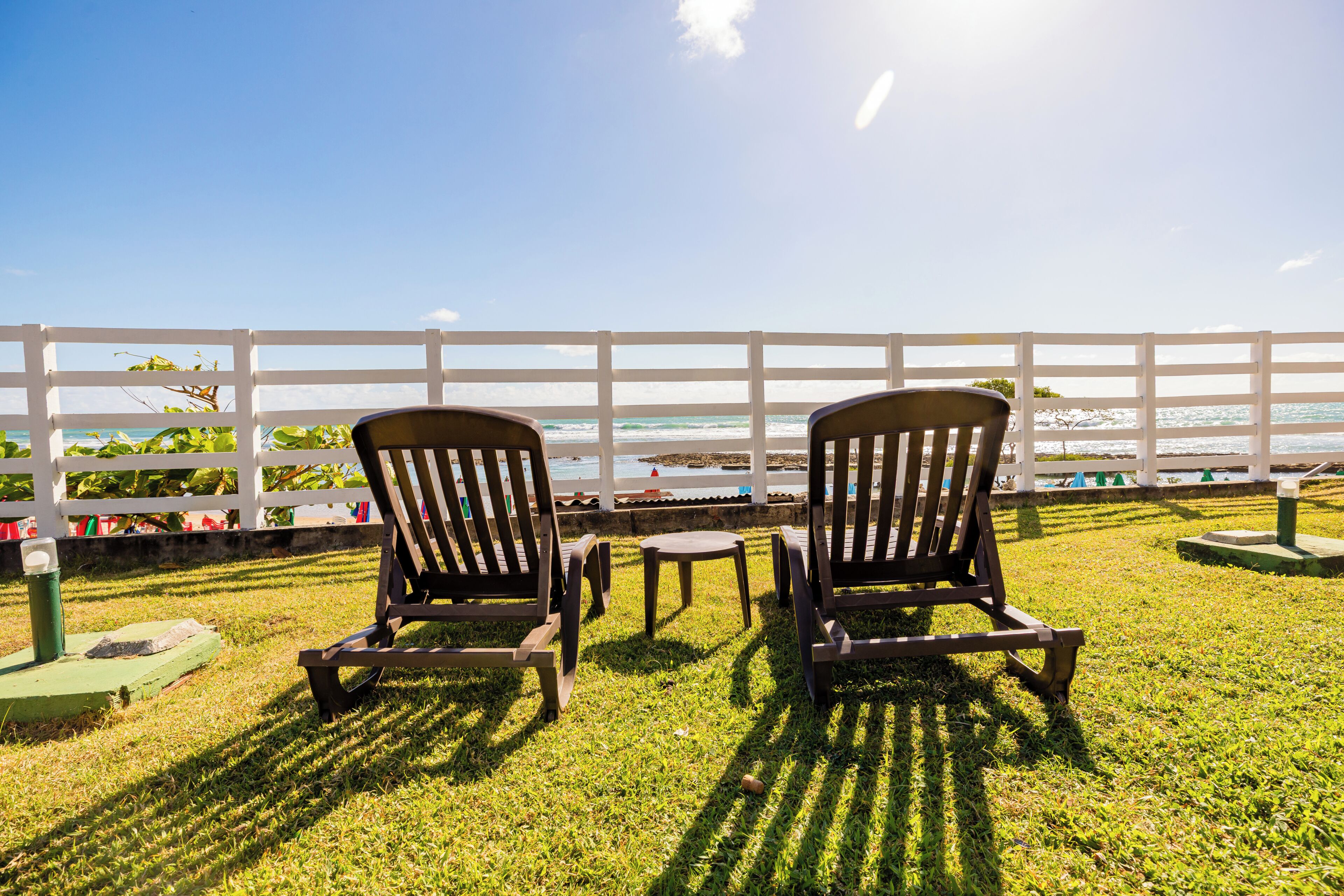 Two wooden chairs facing a white fence overlooking a beach with waves.