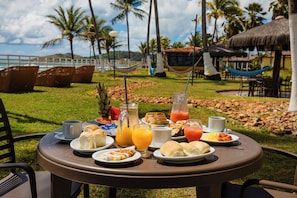 Une table de petit-déjeuner avec divers aliments et boissons, entourée de sièges extérieurs et de palmiers.