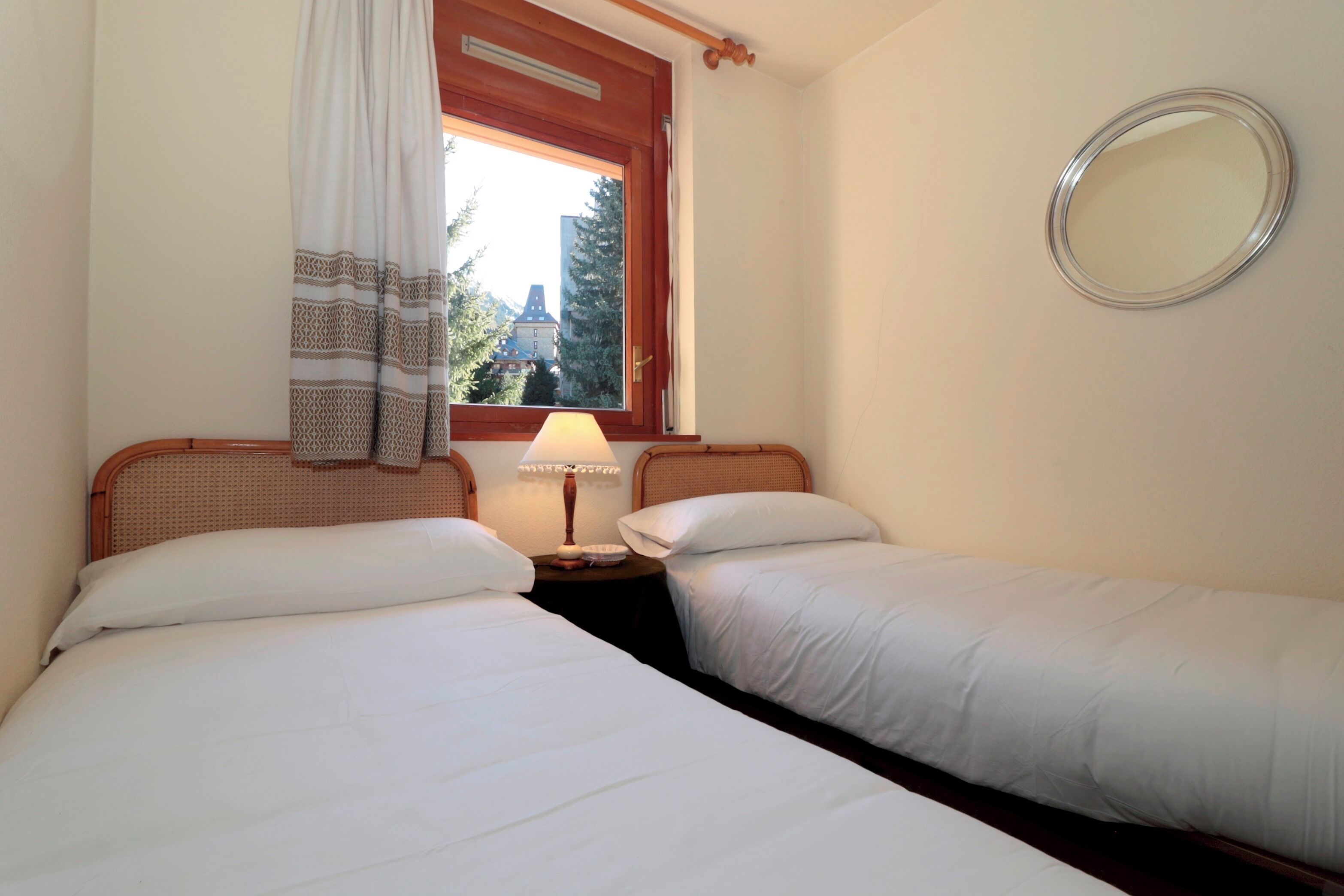 A hotel room with two single beds, a bedside table with a lamp, a window with a view of buildings, and a round mirror on the wall.