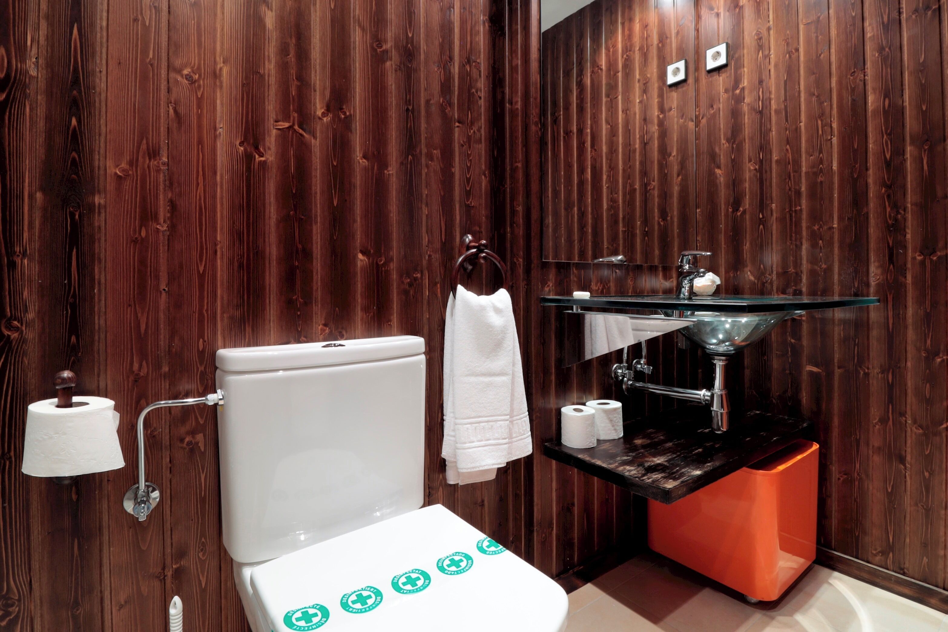 Une salle de bain avec un mur en bois, des toilettes blanches, un lavabo en verre et une poubelle orange.