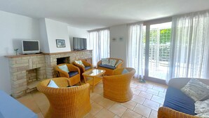A living room with wicker furniture, a fireplace, a television, and large windows. - VILLA NORTH 321B - A2 6+0 - B03215 (Bibione Pineda)
