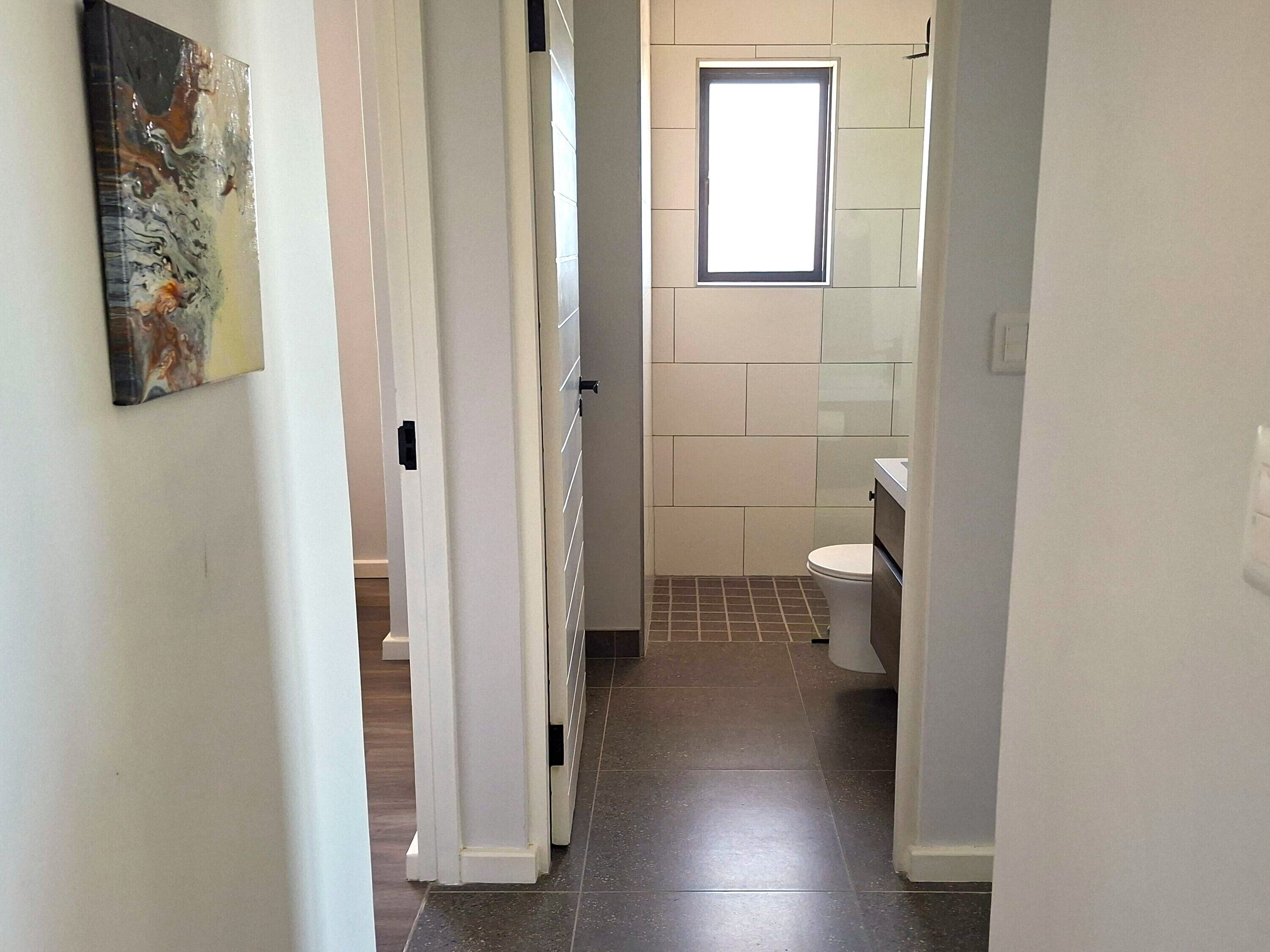 Um corredor estreito que dá para um banheiro com vaso sanitário, janela e piso de azulejos.