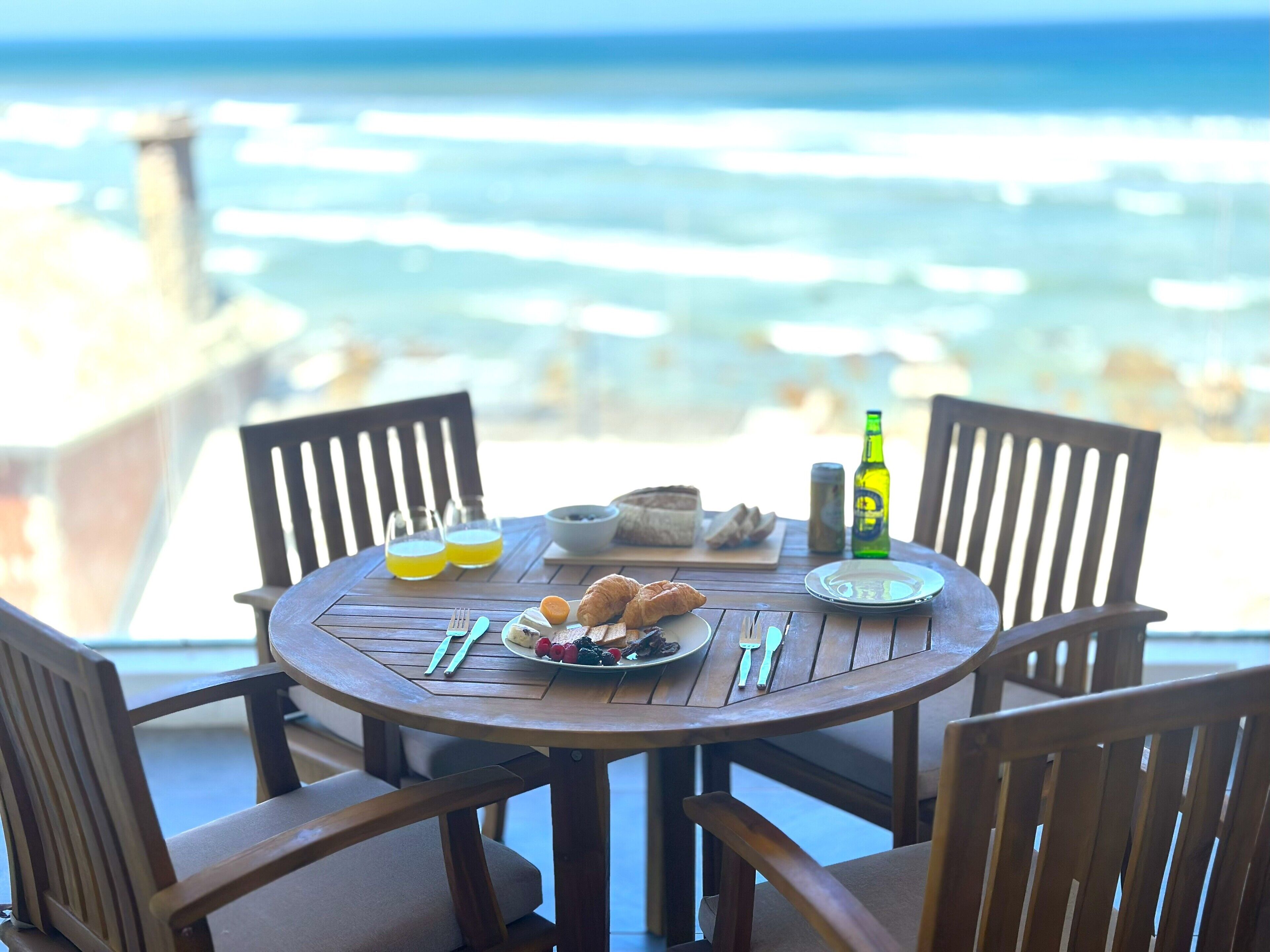 Una mesa redonda de madera con un desayuno, incluyendo croissants, bayas y una botella de cerveza, con vista a una playa.
