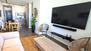 Un salon moderne doté d’un grand téléviseur, d’un canapé, d’une table basse et d’un coin repas avec des chaises et une plante.
