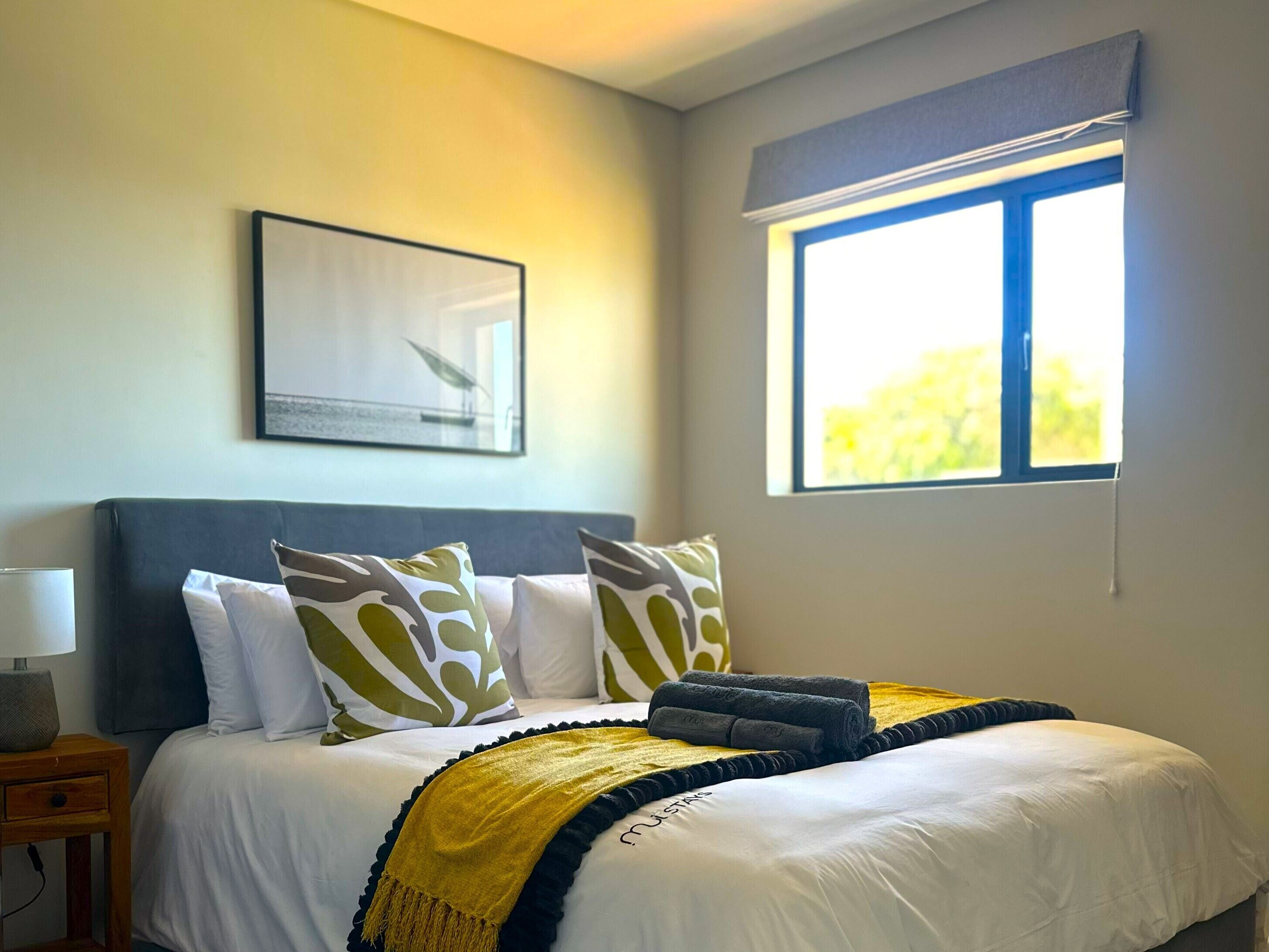 A neatly made bed with a yellow throw blanket, white pillows, and a framed picture on the wall.