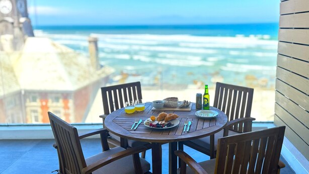 Une table ronde en bois, entourée de chaises, avec une bouteille et des assiettes remplies de nourriture, donnant sur une plage où l’on aperçoit des vagues et, au loin, un clocher.