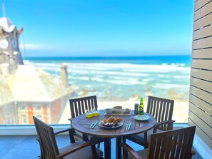 A round wooden table with chairs, a bottle, and plates of food, overlooking a beach with waves and a clock tower in the background. - Wavescapes RipCurl Stay (Muizenberg)