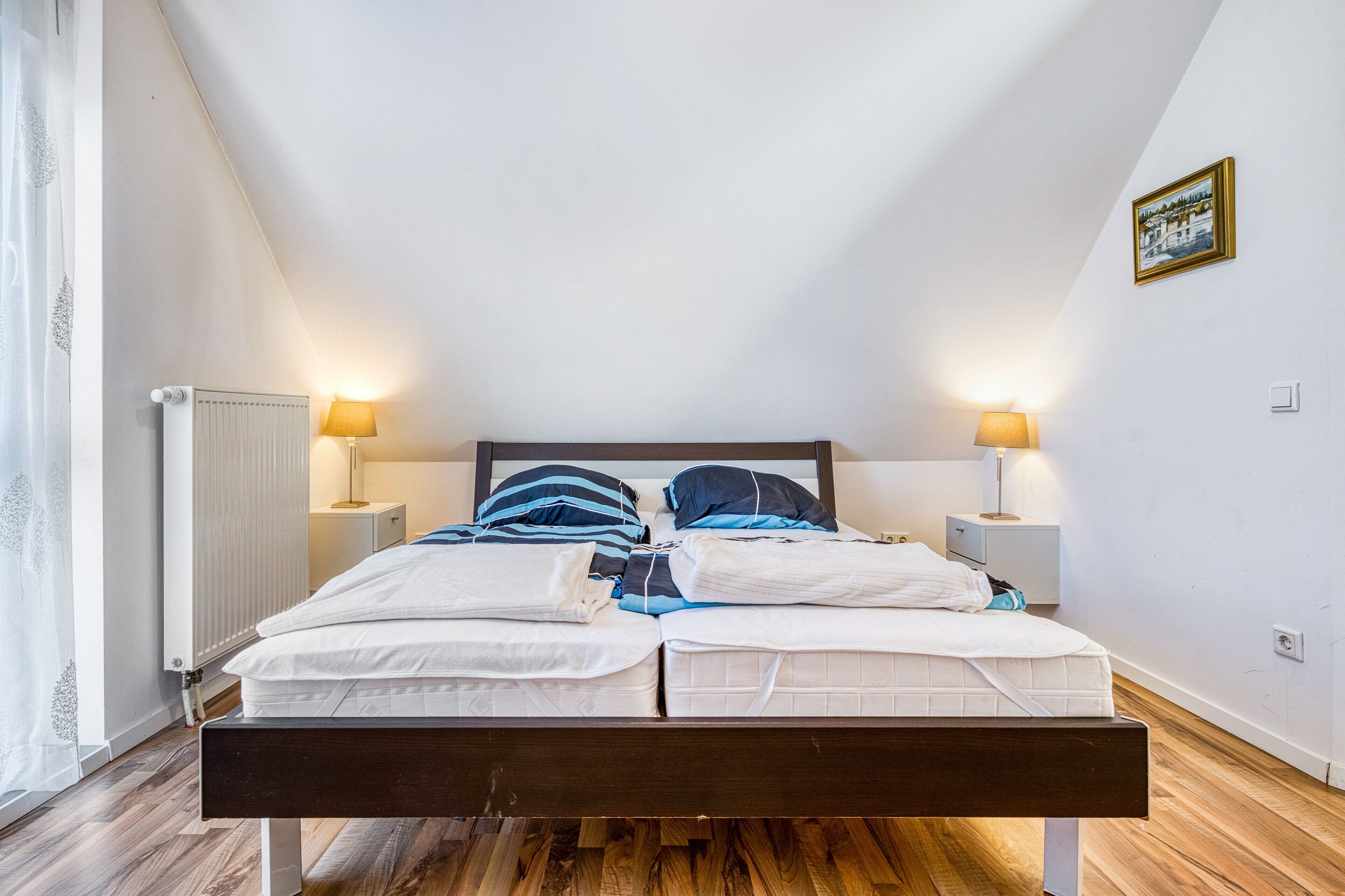 Een slaapkamer met een houten bed, twee nachtkastjes met lampen, een radiator en een schilderij aan de muur.