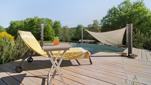 Terraza de madera con piscina, una tumbona cubierta y una mesa con una bebida.