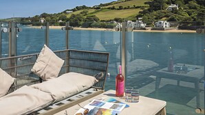A glass-enclosed deck with a sofa and table, overlooking a body of water and a hilly landscape. - Villa 8, Estura (Salcombe)