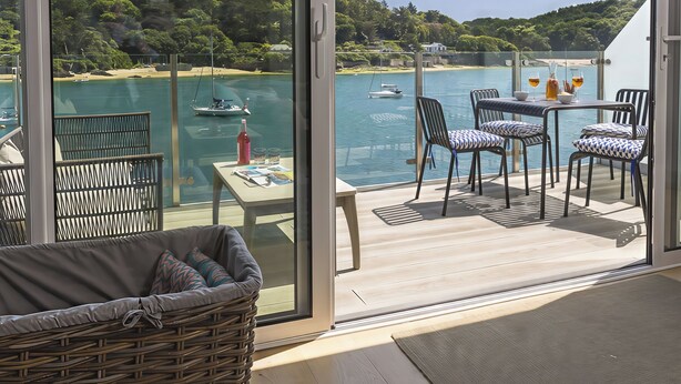 A balcony with a view of a lake, a table with chairs, and a wicker chair. - Villa 8, Estura (Salcombe)