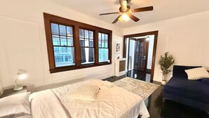 Quarto com duas camas, um ventilador de teto, janela com persianas, um sofá de cor escura e uma mesinha de cabeceira com abajur.