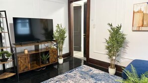 Uma sala de estar com televisão de tela plana sobre um móvel de madeira, um tapete azul e branco e duas plantas em vasos.