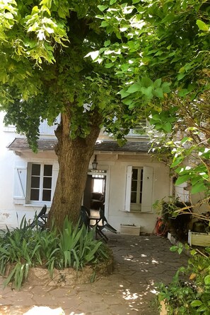 A rustic building with a large tree in front, a stone pathway, and a small outdoor seating area. - House with courtyard and garden (Marolles-sur-Seine)