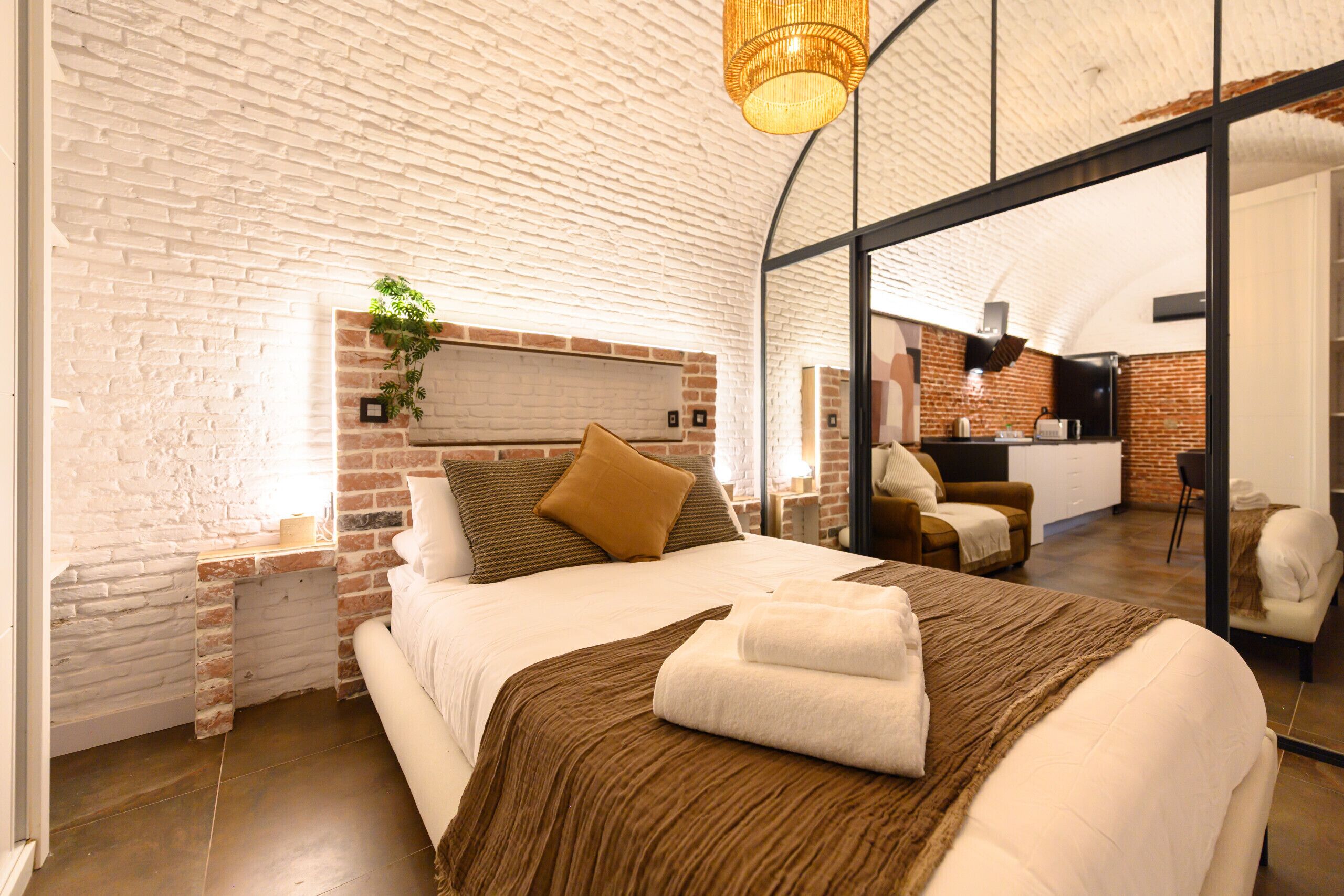 Un dormitorio moderno con pared de ladrillo, una cama grande con cabecera marrón, una planta colgante y vista hacia la zona de la cocina.