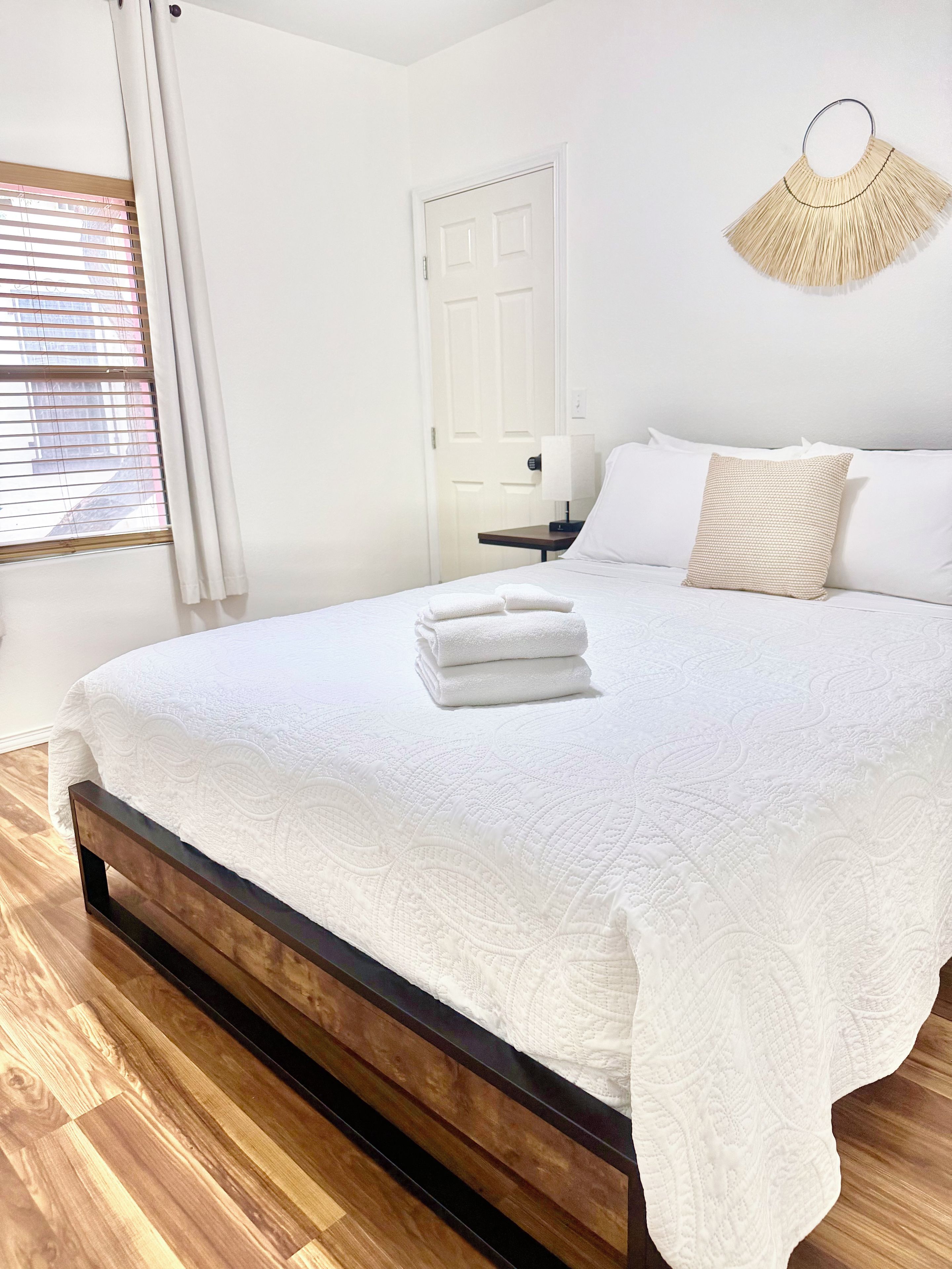 Una cama bien hecha con edredón blanco, una mesita de noche con lámpara y una puerta al fondo.