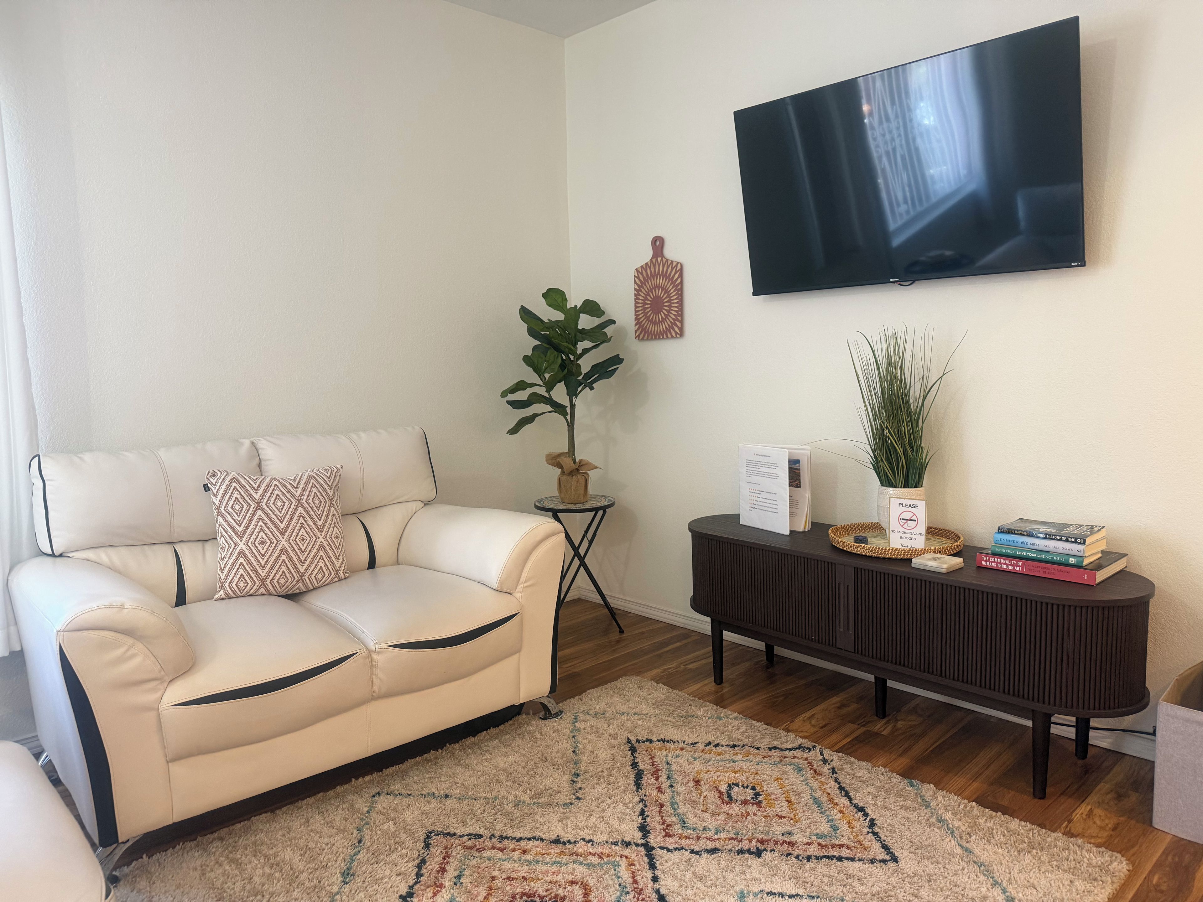 A living room with a beige sofa, a TV mounted on the wall, a small side table with a plant, and a dark console table with books and decorative items.