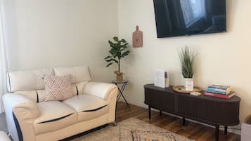 A living room with a beige sofa, a TV mounted on the wall, a small side table with a plant, and a dark console table with books and decorative items.
