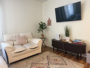 A living room with a beige sofa, a TV mounted on the wall, a small side table with a plant, and a dark console table with books and decorative items. - Pergola Retreat/Prime Central Location (El Paso)