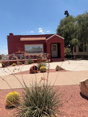 A building with a red exterior, a large window, and a door. There are cacti and desert plants in the foreground. - Pergola Retreat/Prime Central Location (El Paso)