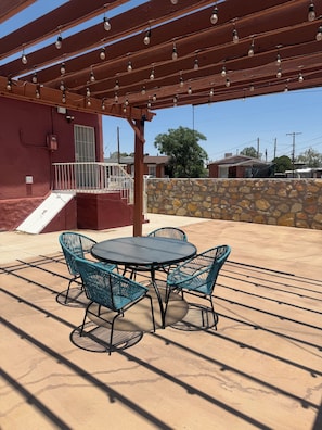 Un patio exterior con una mesa redonda y cuatro sillas, una pérgola con luces colgantes y un muro de piedra.