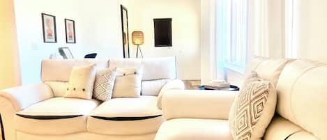 A modern living room with a white leather sofa, patterned cushions, a floor lamp, and a side table with books.