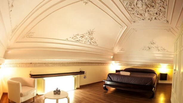 Ein Hotelzimmer mit einem Bett, einem Sessel, einem kleinen Tisch und einer Decke mit aufwendigen Verzierungen.