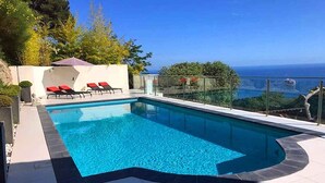 Piscina retangular com água azul cristalina, rodeada por um deck ladrilhado. Há espreguiçadeiras vermelhas e um pequeno guarda-sol. A piscina fica adjacente a um parapeito de vidro com vista panorâmica para o mar e vegetação.