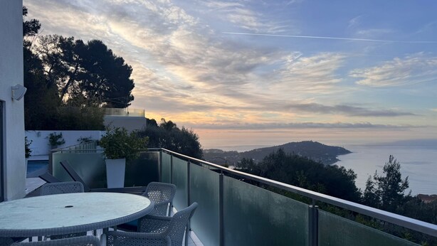Uma varanda com uma mesa redonda e cadeiras de vime, com vista para uma paisagem costeira com árvores e o mar ao longe.