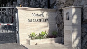Uma entrada de pedra com um portão de metal, uma placa com a inscrição "DOMAINE DU CASTELLET" e um pequeno vaso com plantas.