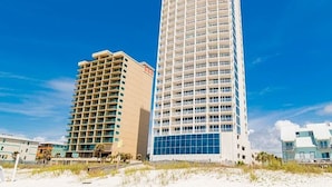 Un grand hôtel à plusieurs étages, doté de balcons, situé près d’une plage de sable où la végétation est clairsemée.