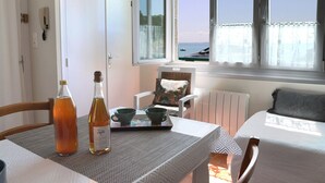 Ein Tisch für zwei ist gedeckt, mit einer Weinflasche und einer Serviertschale mit Tassen und Untertassen. Durch das Fenster ist der Meerblick zu sehen.
