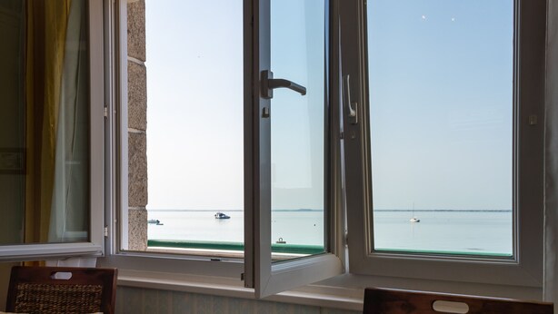 Ein Zimmer mit Meerblick durch ein offenes Fenster, ein Holzbstuhl und ein Tisch mit einem Buch.