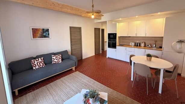 Ein modernes Wohnzimmer mit grauem Sofa, einem Esstisch mit Stühlen sowie einer Küchenzeile mit weißen Schränken und Geräten.
