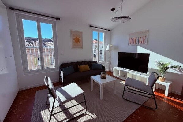 A modern living room with a sofa, coffee table, and chairs, a flat-screen TV, and large windows offering a view of buildings outside. - Charming, bright T3/T4 in the heart of Provence, a stone's throw from everything. (Aubagne)