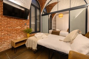 Une chambre moderne avec un mur en briques, un téléviseur à écran plat, un lit avec du linge de maison blanc, un banc en bois et une grande fenêtre.